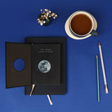 Black notebook with moon illustration on a blue background, accompanied by a cup of tea and pencils.