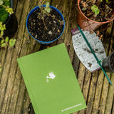 Green gardening notebook on a wooden surface with plants and gardening tools.