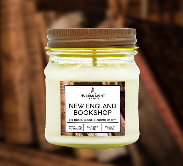 a candle in a clear glass mason jar with off white wax and a label that reads "New England Bookshop" against a soft image of a bookcase