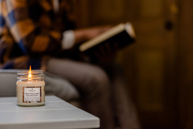 a New England Bookshop candle burning on a side table next to someone seated and reading
