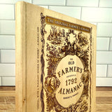 Wooden book style cutting board with cover art from 'The Old Farmer's Almanac' design on a tiled wall background
