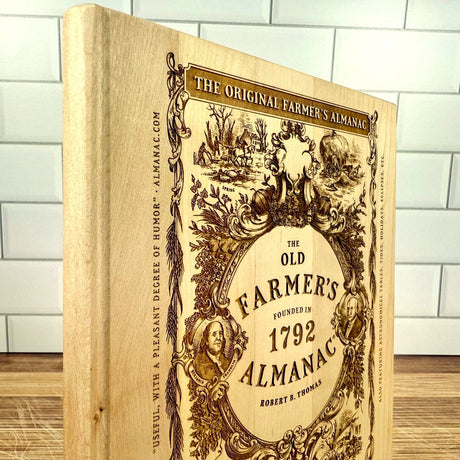 Wooden book style cutting board with cover art from 'The Old Farmer's Almanac' design on a tiled wall background