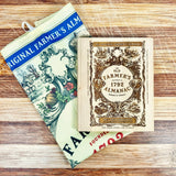 Vintage-style 'Old Farmer's Almanac' cutting board on a wooden surface with a folded Old Farmer's Almanac Tea Towel folded underneath it