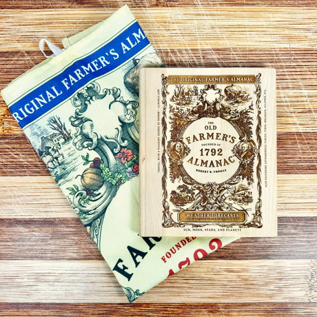 Vintage-style 'Old Farmer's Almanac' cutting board on a wooden surface with a folded Old Farmer's Almanac Tea Towel folded underneath it