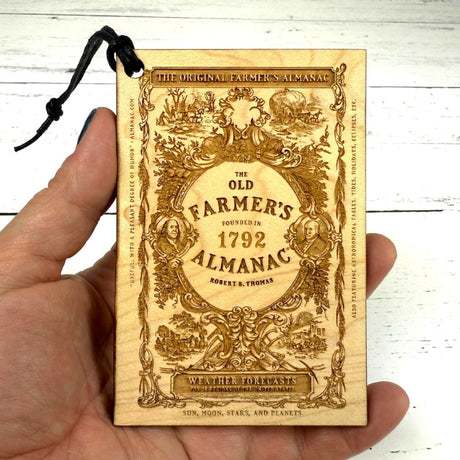 Wooden ornament with 'The Old Farmer's Almanac' design held by a hand on a white wooden surface.