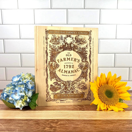 Vintage-style cutting board with 'The Old Farmer's Almanac' cover art on a wooden surface with flowers.