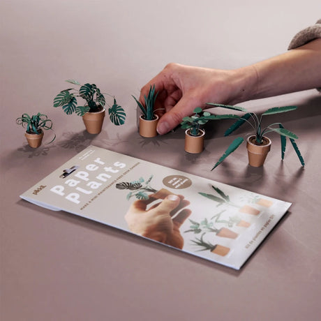 Hand arranging small potted plants next to a 'Paper Plants' book on a table.