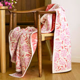 Floral-patterned blanket draped over a wooden chair with a wooden table and flowers in the background.