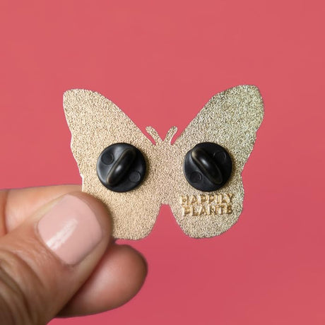 back of Butterfly-shaped enamel pin with 2 rubber backers and 'Happily Plants' text on a pink background