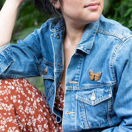 Person wearing a denim jacket with a butterfly pin, standing outdoors.