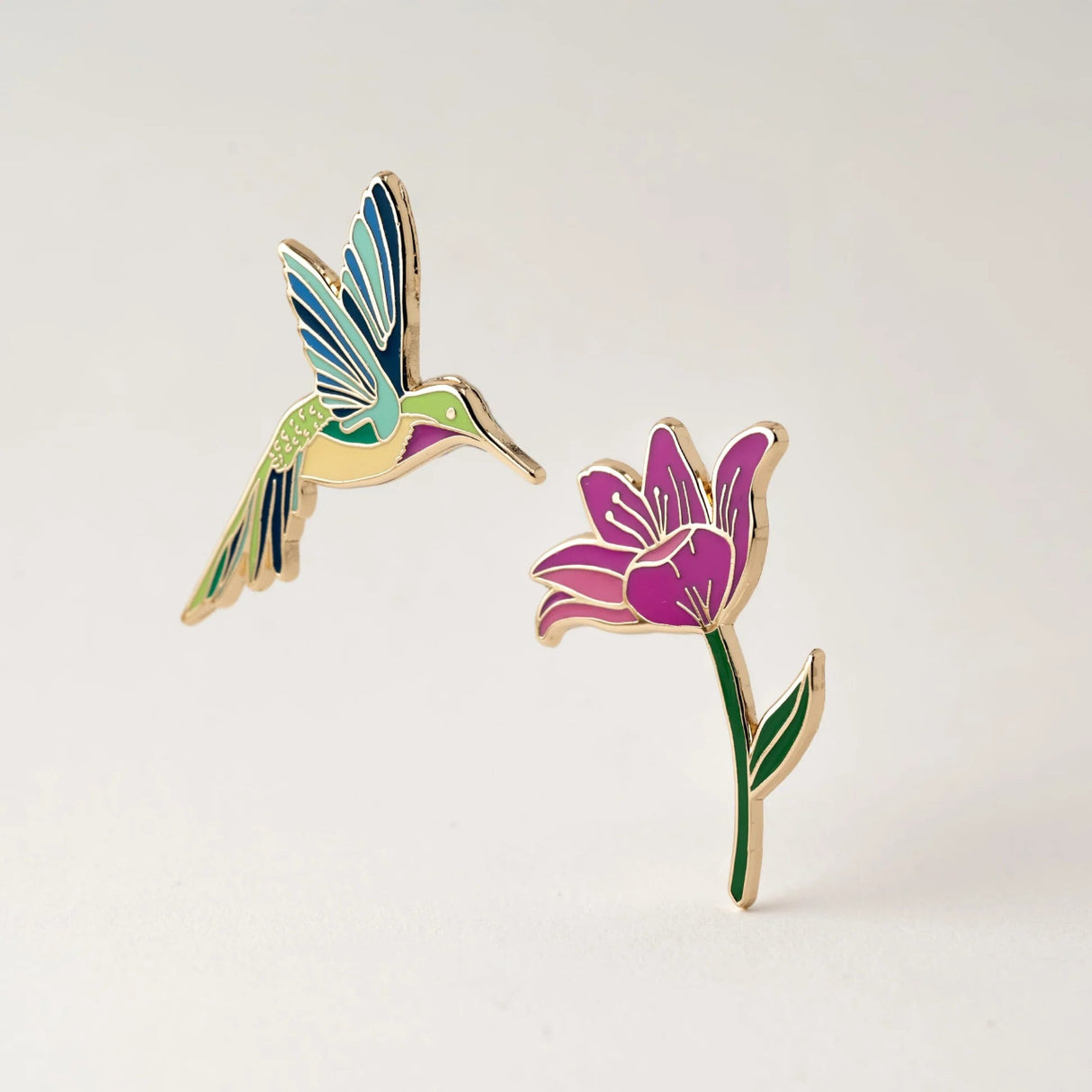 Colorful hummingbird and flower enamel pins on a light gray background
