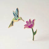 Colorful hummingbird and flower enamel pins on a light gray background