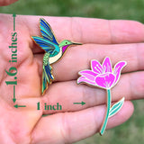 Two enamel pins, one hummingbird and one flower, held in a hand with measurements indicated.