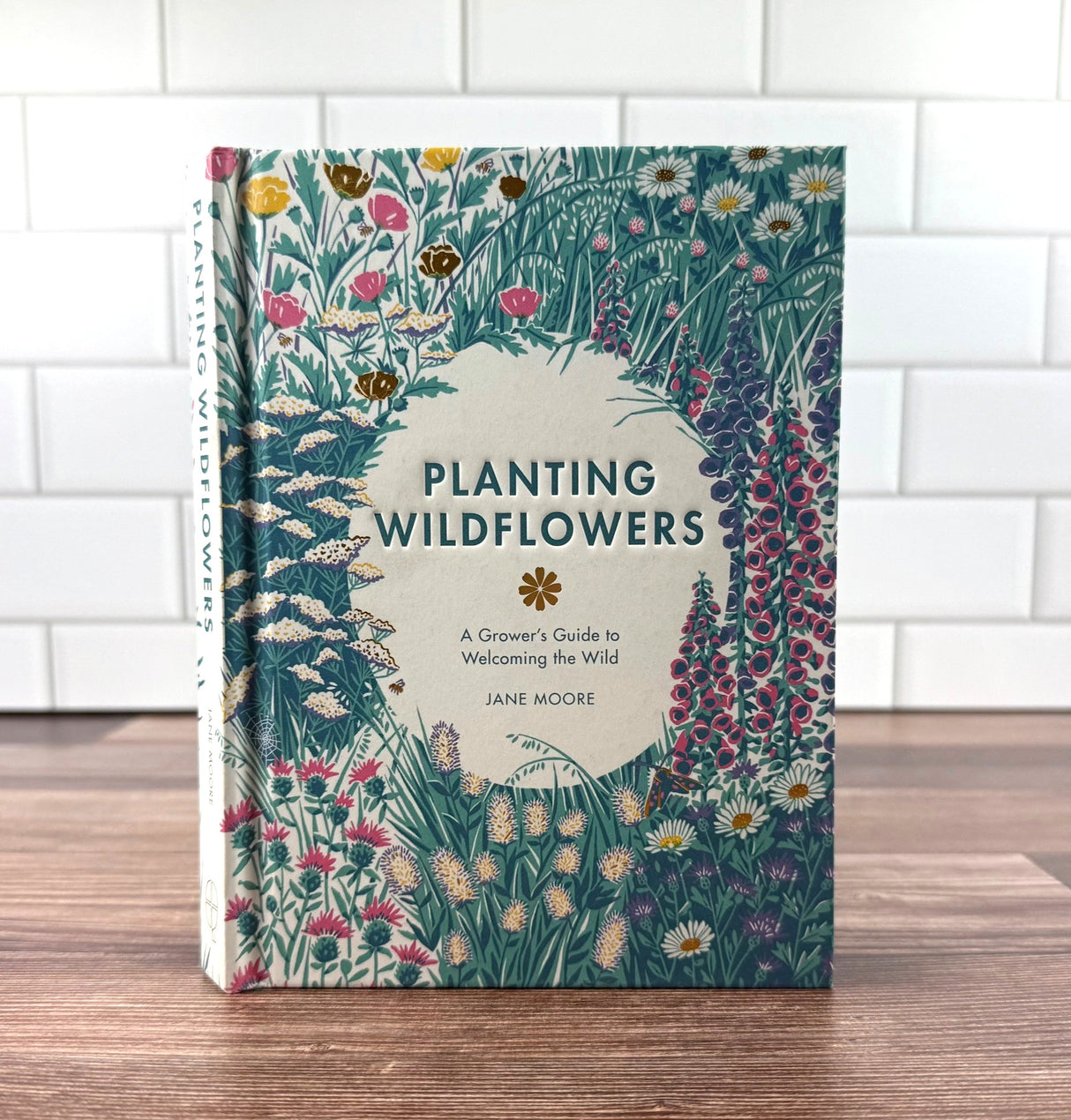 Book titled 'Planting Wildflowers' by Jane Moore on a wooden surface with a white tiled wall background.