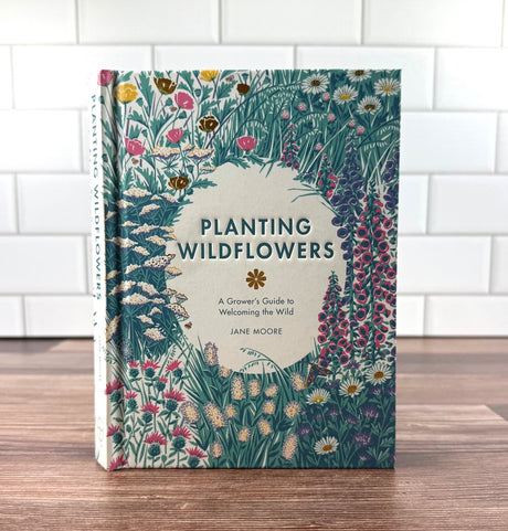 Book titled 'Planting Wildflowers' by Jane Moore on a wooden surface with a white tiled wall background.