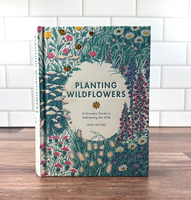 Book titled 'Planting Wildflowers' by Jane Moore on a wooden surface with a white tiled wall background.