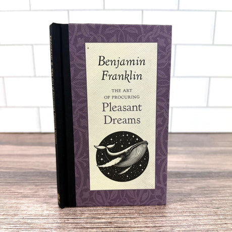 Book titled 'The Art of Procuring Pleasant Dreams' by Benjamin Franklin on a wooden surface with a white tiled wall background.