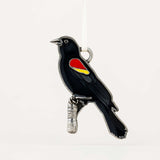 A black bird ornament with a red and yellow wing patch, perched on a small silver branch, photographed against a plain white background.