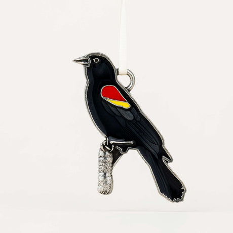 A black bird ornament with a red and yellow wing patch, perched on a small silver branch, photographed against a plain white background.