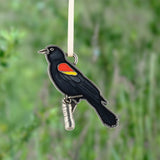 A black bird ornament with a red and yellow wing patch, hanging from a white ribbon against a blurred green outdoor background.