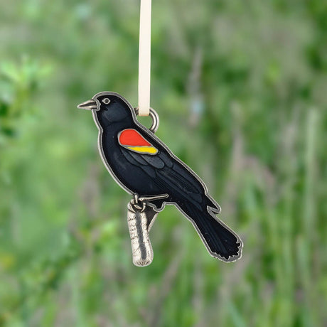 A black bird ornament with a red and yellow wing patch, hanging from a white ribbon against a blurred green outdoor background.