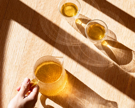 Hand holding a glass of yellow liquid on a wooden surface with two additional glasses in the background.