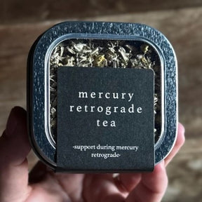 a close up of the mercury retrograde tea tin with elegant black label and clear top to show the loose leaf tea inside