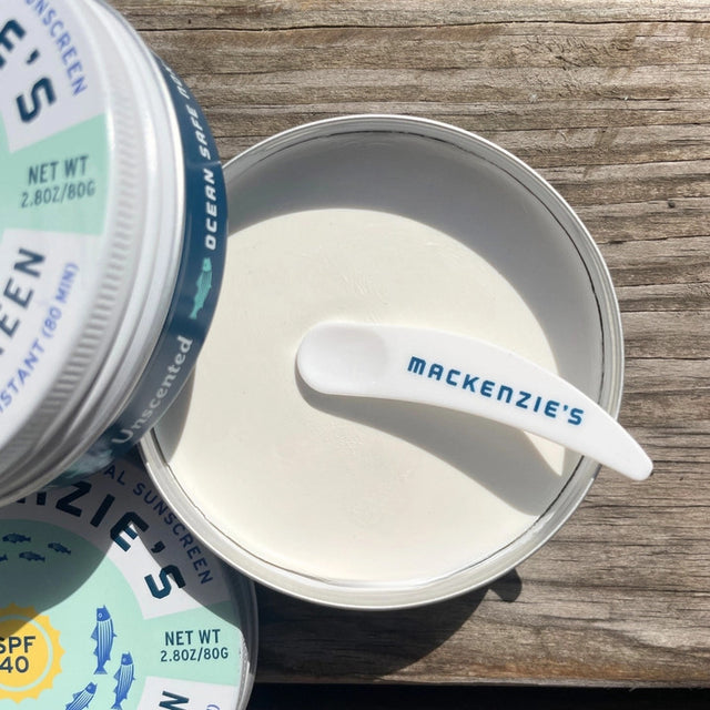 tins of ocean safe sunscreen on a rustic dock - one of the tins is open showing the plastic applicator scoop and the creamy white sunscreen