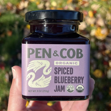 Jar of Pen & Cob Organic Spiced Blueberry Jam held in a hand with a blurred natural background