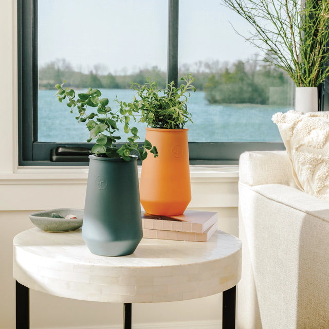 Two potted plants on a round marble table with a window view of water and trees.