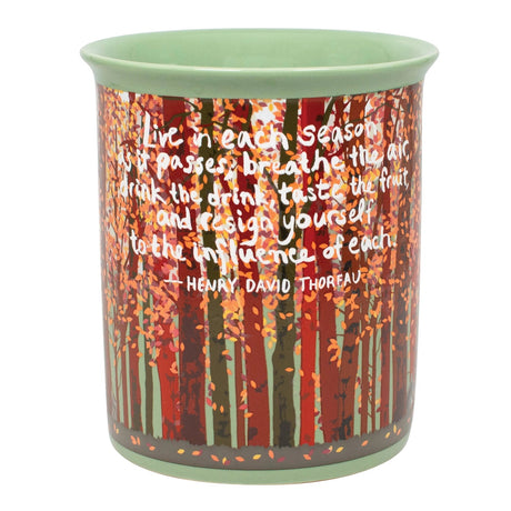 A ceramic mug featuring a forest design with red and orange autumn leaves and white text of a famous quote by Henry David Thoreau, with a light green interior and rim.