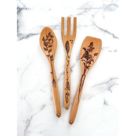 Set of three wooden utensils with engraved vegetable designs on a marble surface - tomato plant, pea pods, and carrot