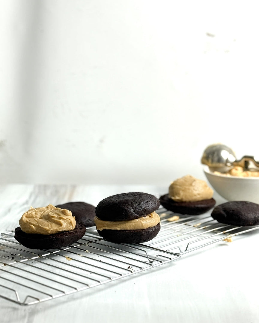 Chocolate cake or cookie whooperas filled with peanut butter cream, arranged on a metal cooling rack with a bowl of more cream in the background.