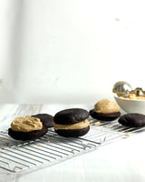 Chocolate cake or cookie whooperas filled with peanut butter cream, arranged on a metal cooling rack with a bowl of more cream in the background.