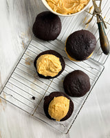 Four chocolate cupcakes, including two topped with swirls of creamy peanut butter frosting, arranged on a wire cooling rack over a white wooden surface. A bowl of peanut butter frosting and a metal spatula are visible in the background.
