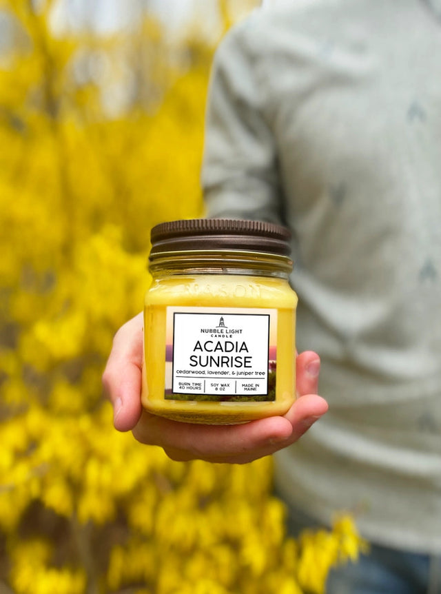 the acadia sunrise candle held in a model's hand with yellow blooming hedges in the background