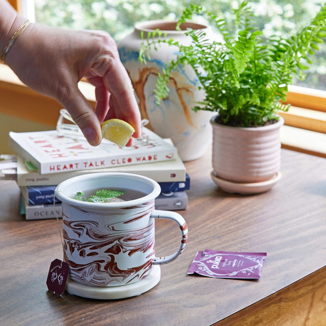 The mostly medicinal lemon balm altered herbs mug on a tabletop next to a potted fern. the mug is filled with steaming tea and some lemon balm leaves with a tea bag package nearby - a hand hovers over the mug squeezing a fresh lemon slice into the tea