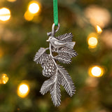 A delicately detailed handcrafted pewter ornament of a bird on a pine bough hanging from a christmas tree from a green silk ribbon