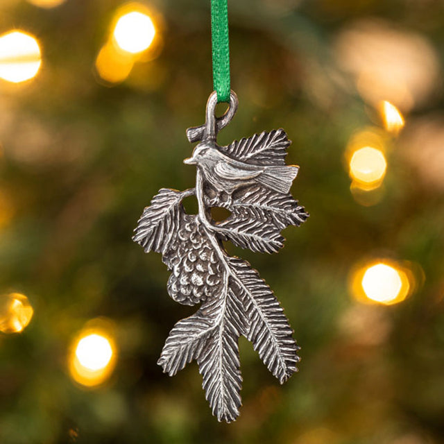 A delicately detailed handcrafted pewter ornament of a bird on a pine bough hanging from a christmas tree from a green silk ribbon