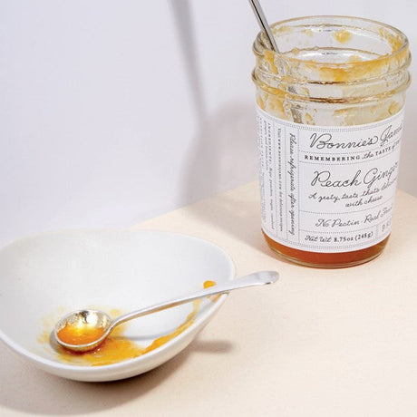 A nearly empty jar of Bonnie's Peach Ginger jam next to a white bowl with a silver spoon coated in jam