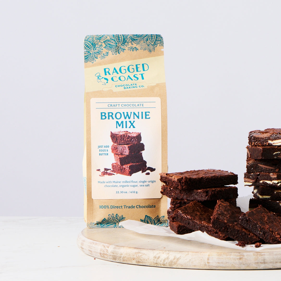 A bag of Ragged Coast Craft Chocolate Brownie Mix stands on a light wood board next to several stacked brownie pieces on a white background.