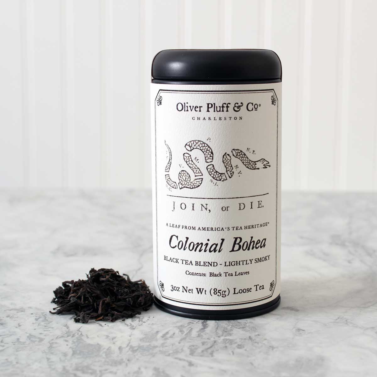 Oliver Pluff & Co Colonial Bohea tea tin with Join or Die political cartoon by Benjamin Franklin on the label. Tea Tin stands on a white marble surface with a heap of loose black tea leaves next to it against a white background