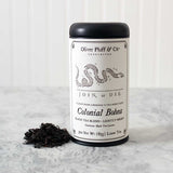 Oliver Pluff & Co Colonial Bohea tea tin with Join or Die political cartoon by Benjamin Franklin on the label. Tea Tin stands on a white marble surface with a heap of loose black tea leaves next to it against a white background