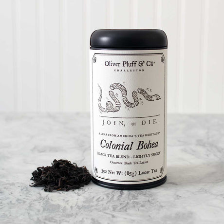 Oliver Pluff & Co Colonial Bohea tea tin with Join or Die political cartoon by Benjamin Franklin on the label. Tea Tin stands on a white marble surface with a heap of loose black tea leaves next to it against a white background