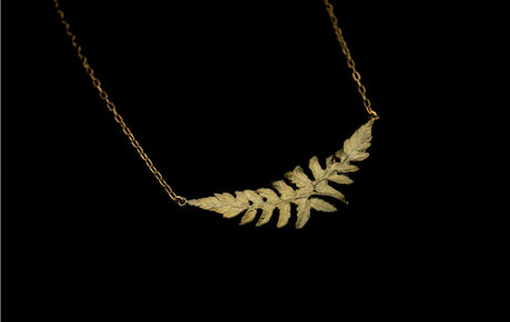 an angled close up view of a delicate chain with a pendant featuring two overlapping fern sprigs cast in bronze with a delicate green patina