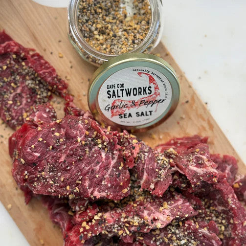 an open jar of garlic and pepper sea salt on a cutting board with red meat that is sprinkled with the garlic and pepper salt