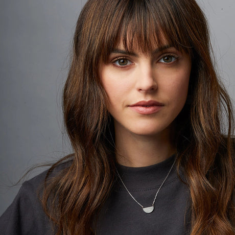 A silver necklace with a half moon pendant seen on a model showing the scale of the necklace and the chain length - the pendant lands just below the collarbone