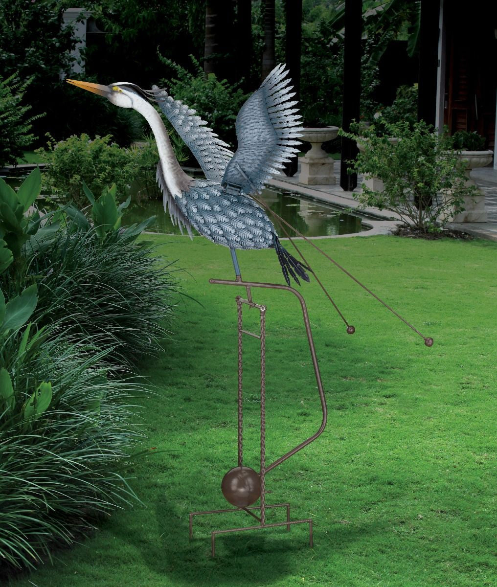 Full view/side angle of a metal heron sculpture in mid-flight, with outstretched wings and an elongated neck installed in a lush, green garden. The sculpture features detailed feather patterns and is mounted on a tall, sturdy frame with a spherical weight and pronged legs at the base. Two rods with weights are attached to the bird's body, enhancing its kinetic movement. The heron is painted in shades of blue and gray, and its long yellow beak is pointed forward.