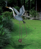 Full view/side angle of a metal heron sculpture in mid-flight, with outstretched wings and an elongated neck installed in a lush, green garden. The sculpture features detailed feather patterns and is mounted on a tall, sturdy frame with a spherical weight and pronged legs at the base. Two rods with weights are attached to the bird's body, enhancing its kinetic movement. The heron is painted in shades of blue and gray, and its long yellow beak is pointed forward.