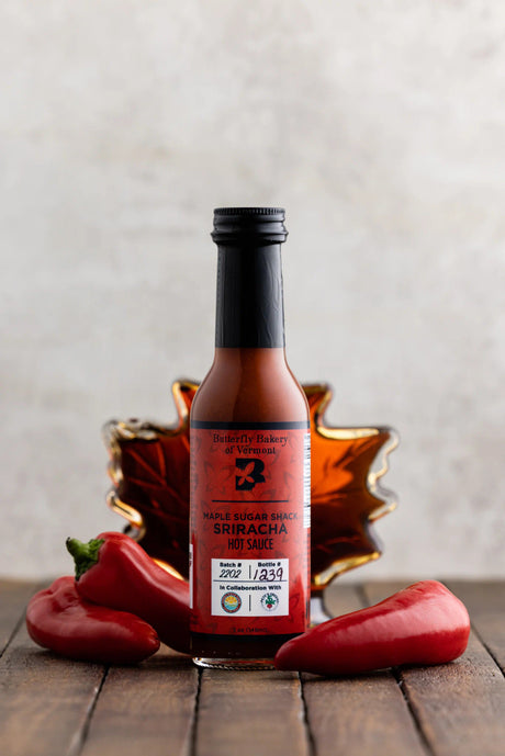 a bottle of maple sugar shack sriracha on a dark wood table surrounded by red serrano and jalepeno peppers with a maple leaf shaped bottle of maple syrup behind it